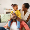 Cheerful little girl sitting on father shoulder while playing with mother at home. Happy black  family enjoying weekend at home. Cute little daughter sitting on fathers shoulder and play with her mom.